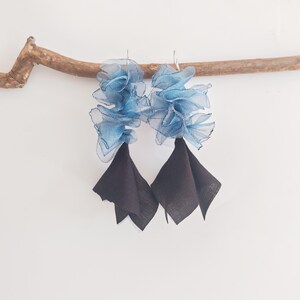 May include: A pair of handmade earrings featuring a layered, ruffled design in shades of blue and black. The top portion of each earring is a cluster of sheer, light blue fabric, while the bottom has a black, square-shaped fabric.