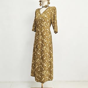 May include: A floral print dress in olive green with a V-neck and three-quarter sleeves is displayed on a mannequin. The dress is accessorized with a straw hat, earrings, and a choker. Sandals with bows are at the base of the mannequin.