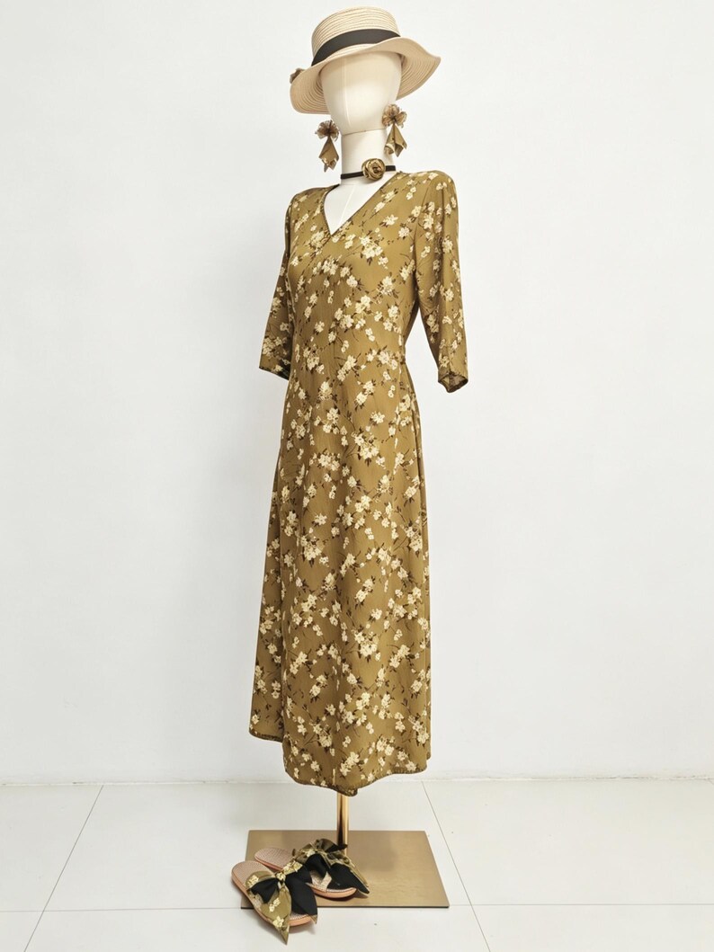 May include: A floral print dress in olive green with a V-neck and three-quarter sleeves is displayed on a mannequin. The dress is accessorized with a straw hat, earrings, and a choker. Sandals with bows are at the base of the mannequin.