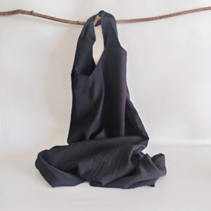 May include: A black tote bag made of a soft, flowing fabric. The bag has a wide opening and a single pocket. It is hanging from a wooden branch against a white background. The bag is designed to be carried over the shoulder.