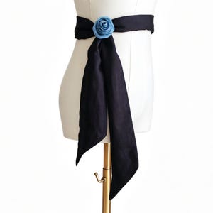 May include: A black sash belt with a light blue rose accent. The sash is draped on a mannequin, showcasing its design. The belt is made of a dark fabric and features a decorative floral element.