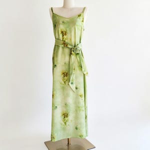 May include: A light green floral print dress with spaghetti straps and a V-neck. The dress features a matching belt with a rose-shaped buckle. The fabric has a watercolor effect with yellow and brown floral accents. The dress is displayed on a mannequin.