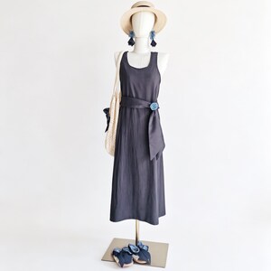 May include: A dark gray sleeveless dress with a matching belt and a blue floral accent. A straw hat, blue earrings, a white tote bag, and blue sandals complete the outfit. The dress is displayed on a mannequin.