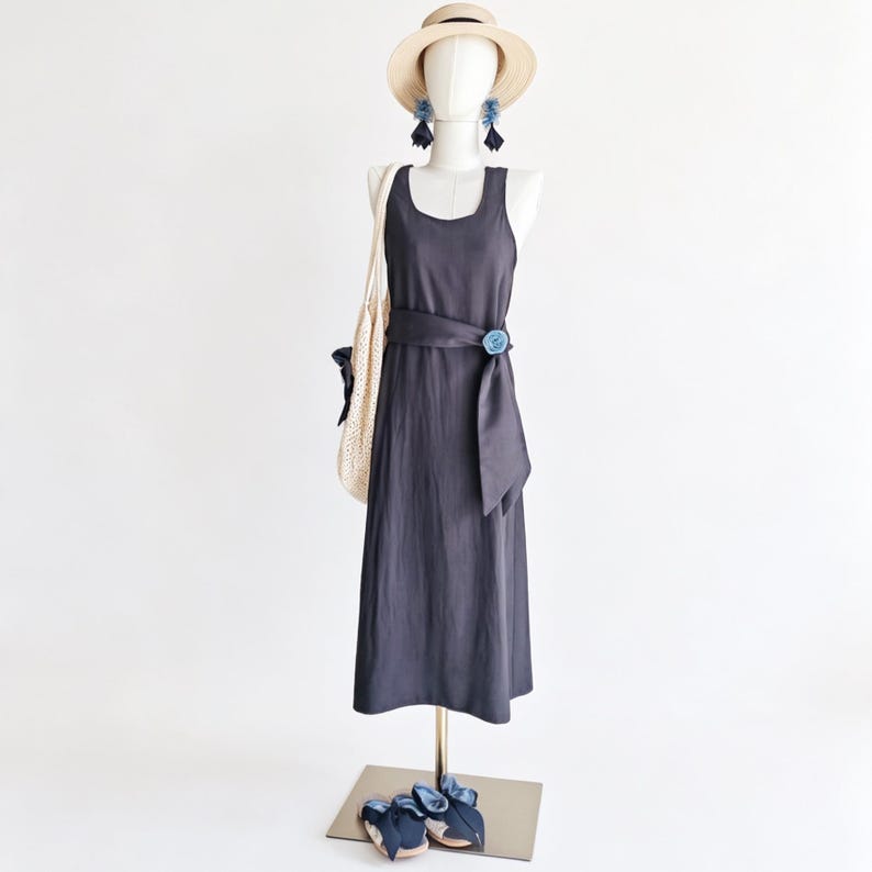 May include: A dark gray sleeveless dress with a matching belt and a blue floral accent. A straw hat, blue earrings, a white tote bag, and blue sandals complete the outfit. The dress is displayed on a mannequin.