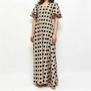 May include: A floor-length beige dress featuring a repeating pattern of large black polka dots, a V-neckline, and short, flared flutter sleeves. The garment has a loose, flowing silhouette that extends to the floor, covering the lower legs.