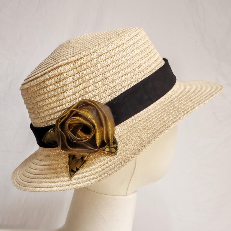 May include: A straw hat with a black band and a gold-colored fabric rose. The hat has a wide brim and a structured crown. The rose is made of a shiny material and is positioned on the side of the hat.