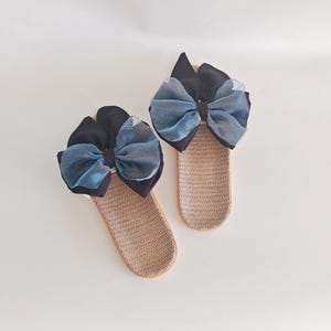 May include: A pair of beige woven slippers, each adorned with a large bow. The bows feature a combination of black and blue fabrics, creating a striking contrast. The slippers are designed for indoor wear.