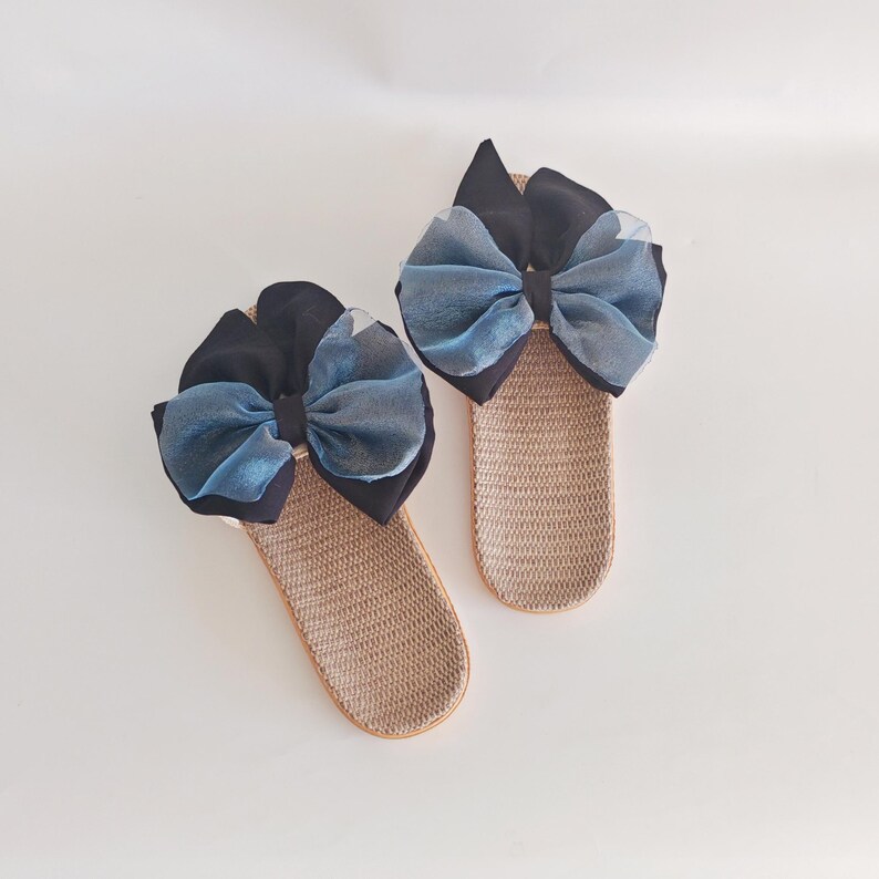 May include: A pair of beige woven slippers, each adorned with a large bow. The bows feature a combination of black and blue fabrics, creating a striking contrast. The slippers are designed for indoor wear.