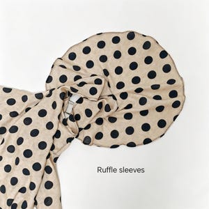 May include: A beige garment featuring a black polka dot pattern and large, circular ruffle sleeves. The fabric has a visible texture, and the edges of the ruffles are finished with a dark stitched hem. The text Ruffle sleeves is visible.
