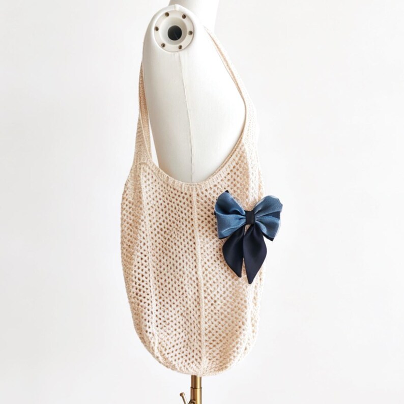 May include: A cream-colored, open-weave tote bag with a single shoulder strap. A large, two-tone blue bow is attached to the front of the bag. The bag is displayed on a mannequin.