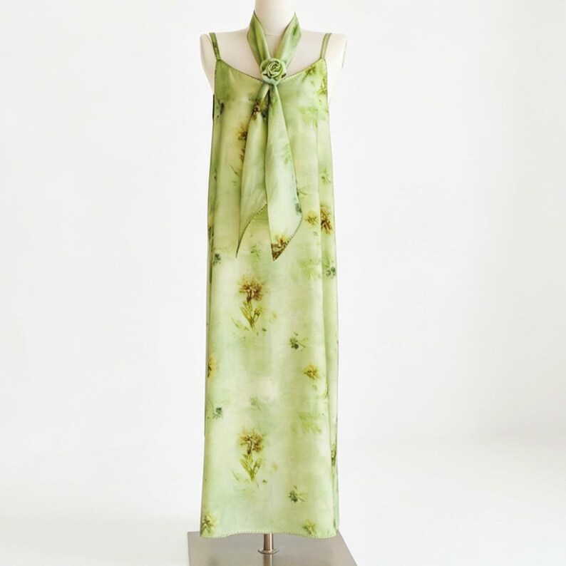 May include: A light green, floor-length dress with spaghetti straps and a matching scarf. The dress features a floral pattern in shades of yellow and green. The scarf is secured with a decorative flower.