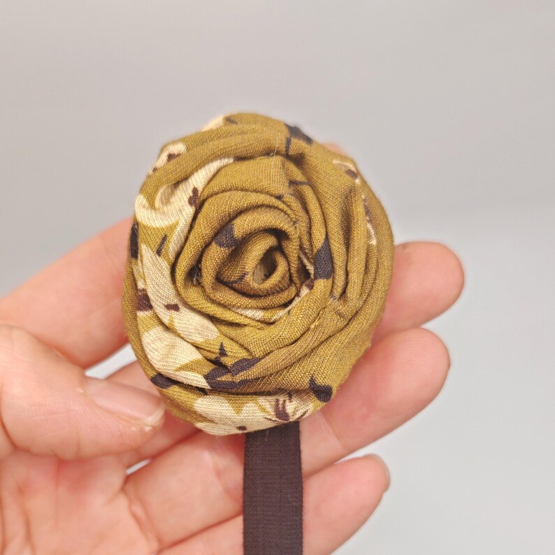 May include: A fabric flower in a rose shape, featuring a mix of olive green, beige, and dark brown patterns. The flower is attached to a dark brown elastic band, likely for use as a hair accessory or embellishment. The design is detailed and textured.