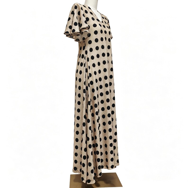 May include: A floor-length beige dress featuring a repeating pattern of large black polka dots. The garment has a V-neckline and short, flared flutter sleeves that extend from the shoulders, creating a loose and flowing silhouette throughout.