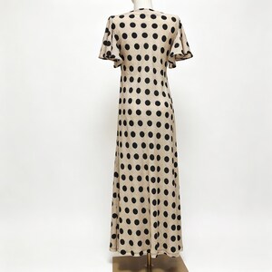 May include: A floor-length dress in a light beige color featuring a repeating pattern of large black polka dots. The garment has short, flared sleeves and a straight silhouette that extends from the shoulders down to the hemline.