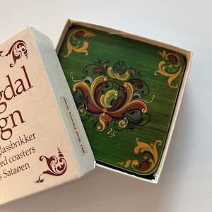 May include: A set of square coasters with a green background and ornate floral designs in shades of yellow, red, and brown. The coasters are in a white box with text in a foreign language.