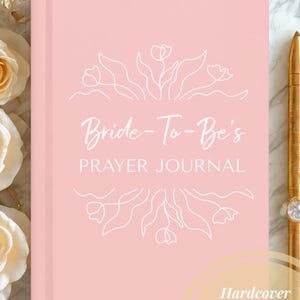 May include: A pink hardcover prayer journal with the text "Bride-To-Be's Prayer Journal" in white script. The cover features a floral design. A gold pen rests on the side. The journal is lined and made of paper.