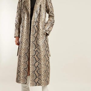 Pode incluir: Um casaco comprido com padrão de pele de cobra em tons de bege, castanho e preto. O casaco tem um design clássico de trench coat com cinto e fenda frontal. A modelo usa botas brancas. Uma peça marcante.