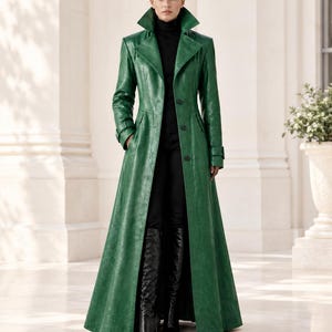 May include: A floor-length emerald green leather trench coat featuring a wide notched collar, long sleeves with buckled cuffs, and a front button closure. The coat is worn over a black turtleneck sweater, black trousers, and black knee-high boots.