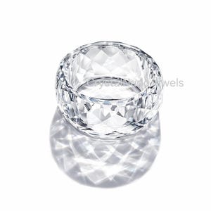 Könnte beinhalten: Ein klarer, facettierter Kristallring mit breitem Band. Der Ring ist rund und hat ein geometrisches Muster aus geschliffenen Facetten. Der Ring wirft einen Schatten auf die weiße Oberfläche darunter. Der Text "crystalnudjewels" ist auf dem Ring sichtbar.