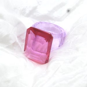 May include: A ring featuring a large, rectangular, faceted hot pink gemstone set atop a translucent, light lavender band. The design showcases a clear, polished finish with sharp, geometric edges and a vibrant color contrast between the pink stone and the purple band.