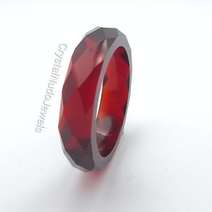 Puede incluir: Un anillo facetado de color rojo intenso. El anillo tiene un diseño geométrico con múltiples superficies planas. El anillo está hecho de un material translúcido, que permite el paso de la luz. El texto "CrystalNudoJewels" está impreso en el lado izquierdo del anillo.