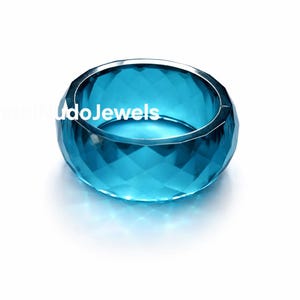 Pode incluir: Um anel de vidro facetado, redondo, de cor turquesa. O anel tem uma faixa larga e um design geométrico. O texto "CrystalNudoJewels" é visível no anel. O anel está sobre um fundo branco.