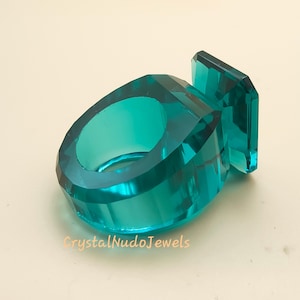 Puede incluir: Un anillo facetado de color turquesa con un acento rectangular. El anillo está hecho de un material transparente y tiene un diseño geométrico. El texto "CrystalNudoJewels" es visible.