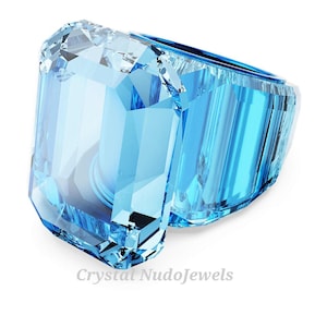 May include: A large, light blue crystal ring with an emerald cut. The ring is faceted, reflecting light. The text "Crystal NudoJewels" is at the bottom of the image.