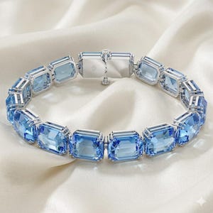 May include: A luxurious bracelet featuring rectangular, light blue gemstones set in a silver-toned metal. The bracelet is displayed on a white, silky fabric, highlighting the sparkle and clarity of the stones. The clasp is also silver-toned.
