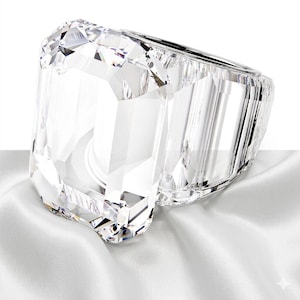 Pode incluir: Um anel de cristal transparente facetado com uma grande pedra retangular de corte esmeralda como peça central. A banda é larga e também construída em cristal transparente facetado, criando uma aparência coesa, transparente e altamente reflexiva.