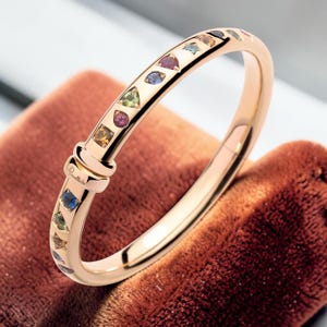 May include: A gold bangle bracelet adorned with a variety of colourful gemstones. The gemstones are in different shapes and colours, including blue, green, red, and yellow. The bracelet has a clasp and is displayed on a brown velvet surface.