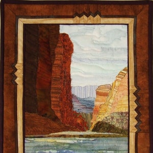 Op de afbeelding: Een textielkunstwerk dat een canyonlandschap uitbeeldt. Het kunstwerk toont een rivier, rode rotsformaties en een blauwe lucht. Het stuk is omlijst in bruin- en beigetinten, met decoratieve hoekaccenten.