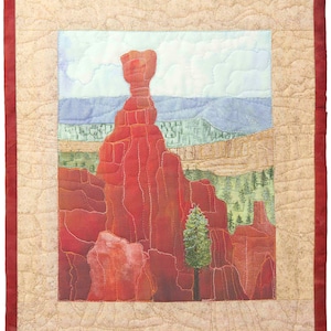 May include: A quilted landscape featuring a red rock formation under a blue sky with white clouds. The artwork includes a green tree and a mountain range in the background. The quilt has a beige border with a red frame.