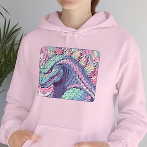 May include: A pale pink hoodie with a Godzilla graphic. The graphic is a colourful illustration of the monster in shades of blue, green, and pink, set against a rainbow background. The hoodie has a kangaroo pocket and a drawstring hood.