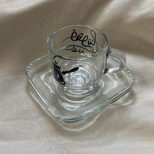 Puede incluir: Un juego de taza y platillo de té de vidrio transparente. La taza tiene un asa y está decorada con escritura negra y un gráfico negro. El platillo es cuadrado con bordes redondeados. El juego está sobre una superficie color crema.
