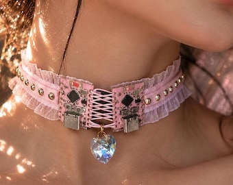 Op de afbeelding: Een lichtroze choker met een geplooide stoffen rand, metalen studs en twee geïntegreerde roze printplaatsegmenten verbonden door roze veters. Een doorzichtige, gefacetteerde hartvormige hanger hangt in het midden van de veters.