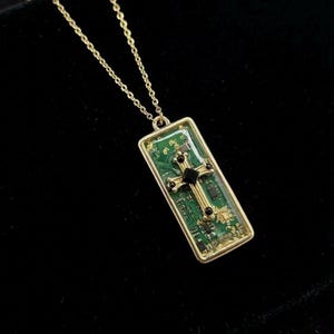 Op de afbeelding: Goudkleurige ketting met een rechthoekige hanger. De hanger heeft een groen printplaatontwerp en een gouden kruis met een zwart midden. De ketting heeft een fijne ketting en staat tegen een zwarte achtergrond.