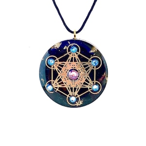 Pode incluir: Um colar com pingente redondo com fundo azul escuro, detalhes dourados e um design geométrico dourado. O pingente apresenta uma gema rosa central e gemas azuis. O colar tem um cordão preto.