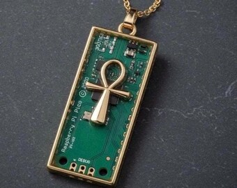 Op de afbeelding: Gouden ketting met een hanger met een groen printplaatontwerp en een gouden ankh-symbool. De rechthoekige hanger heeft de tekst "Raspberry Pi Pico © 2020" en "DEBUG". De ketting is ook goudkleurig. De hanger is ongeveer 4 cm lang.