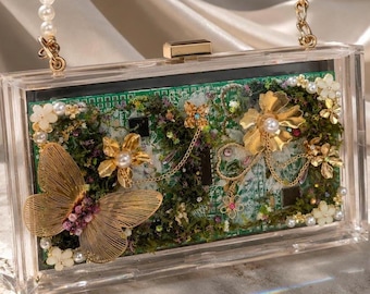Op de afbeelding: Een rechthoekige clutch van doorzichtig acryl met een groene printplaat aan de binnenkant, versierd met goudkleurige metalen vlinders en bloemen, kleine witte kunstparels en groen mos, afgewerkt met een parelmoeren handvat.