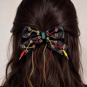 Peut inclure: Un nœud pour cheveux noir doté d'œillets métalliques argentés lacés avec des cordons fins rouges et jaunes. Les cordons s'étendent depuis le nœud, avec un connecteur jack audio rouge et un jaune aux extrémités. Une petite pièce rectangulaire verte est au centre.