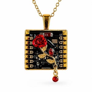 Puede incluir: Un collar de tono dorado con un colgante cuadrado hecho de una placa de circuito negra. El colgante está enmarcado en oro con un borde tachonado y muestra un adorno de rosa roja con hojas doradas y un dije de cristal rojo colgante.
