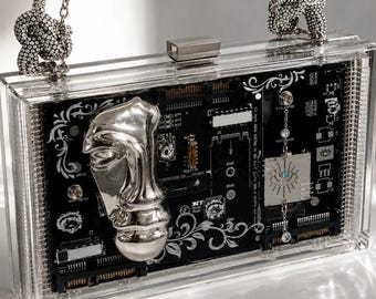 Op de afbeelding: Een rechthoekige clutch van doorzichtig acryl met een zwarte printplaat aan de binnenkant en witte filigraanpatronen. Een groot zilverkleurig metalen gezichtssculptuur is aan de voorzijde bevestigd, naast een zilveren ketting met een blauw oog.