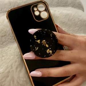 May include: Black smartphone with a gold-coloured frame and a black pop socket. The pop socket features gold leaf accents. The phone is held in a hand with light pink nail polish.
