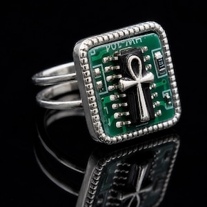Könnte beinhalten: Ein silberfarbener Ring mit quadratischer Fassung und einer grünen Leiterplatteneinlage. In der Mitte der Leiterplatte befindet sich ein schwarzes rechteckiges Bauteil, das mit einem silbernen Ankh-Symbol verziert ist. Der Ring hat eine doppelte Ringschiene.