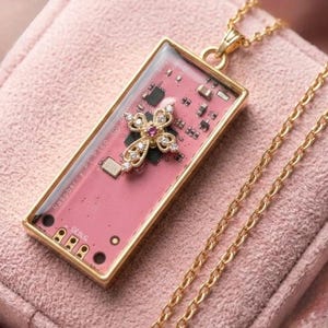 May include: Gold-toned necklace featuring a rectangular pendant with a pink circuit board design. A jeweled cross with a central pink stone and clear accents adorns the pendant. The text "Raspberry Pi Pico ©" and "DEBUG" are visible. The chain is also gold.