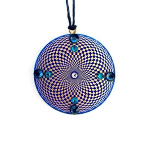 Pode incluir: Um pingente redondo azul com um padrão xadrez dourado e azul. O pingente é adornado com várias pedras preciosas azuis e verde-azuladas. Um cordão preto é preso na parte superior com um fecho dourado.