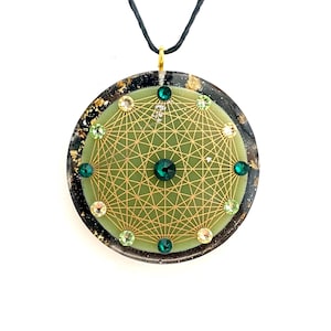 Pode incluir: Um colar com pingente redondo com um cordão preto. O pingente apresenta um centro verde claro com um design geométrico dourado e é adornado com cristais verdes e transparentes. A borda externa é preta com flocos dourados.
