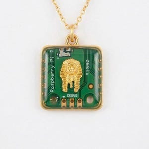 Puede incluir: Collar dorado con un colgante cuadrado. El colgante presenta un diseño de placa de circuito verde, una figura de faraón dorada y los textos "Raspberry Pi P" y "DEBUG".
