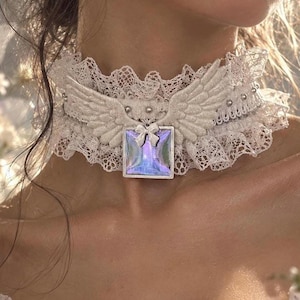 May include: A white lace choker featuring a central square iridescent pendant with a small bow accent. The choker is adorned with glittery white angel wings on either side and small silver studs along the lace band, creating a layered texture.